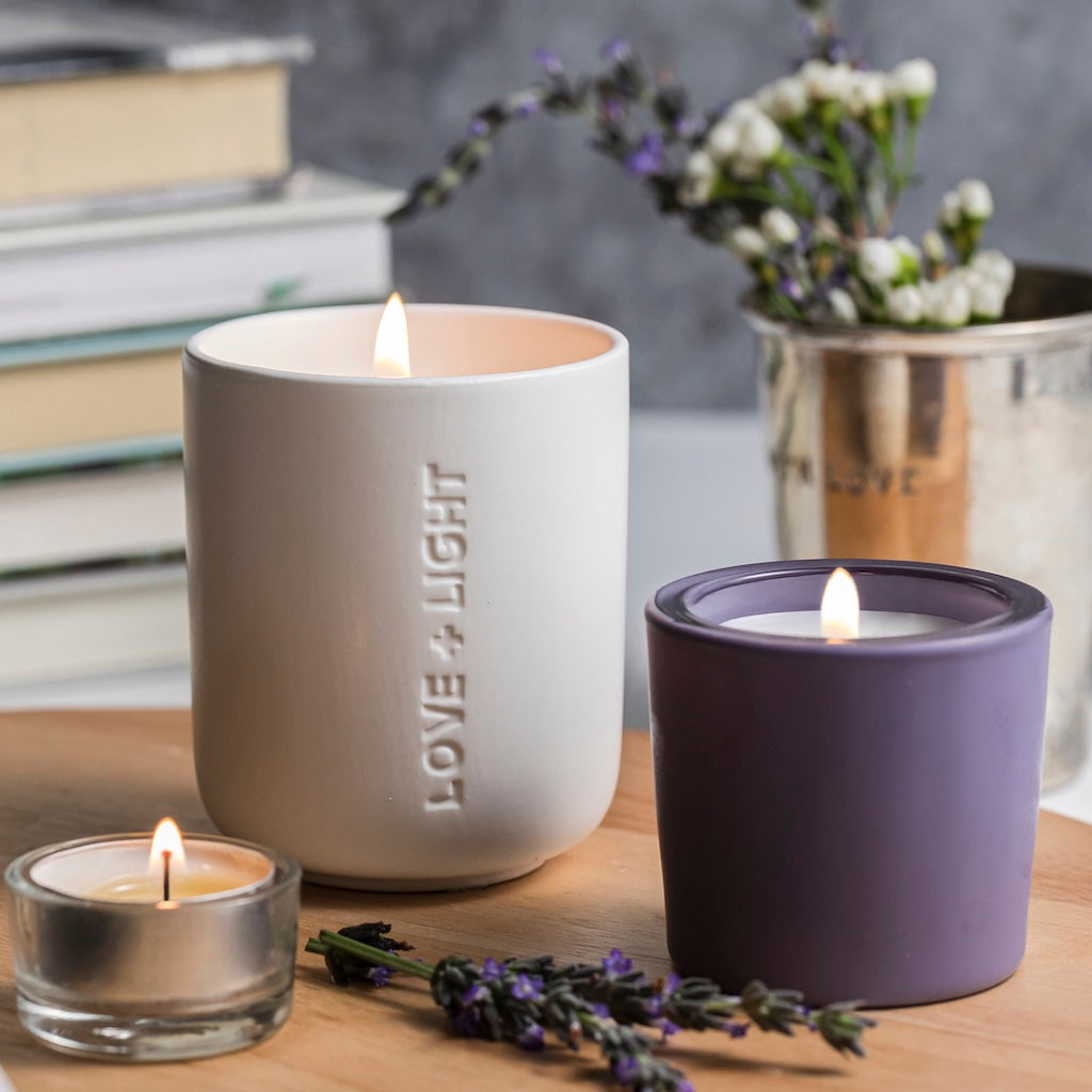 Candle in a white container with 'LOVE + LIGHT' text, surrounded by a purple votive, tea light, books, and flowers on a wooden surface.