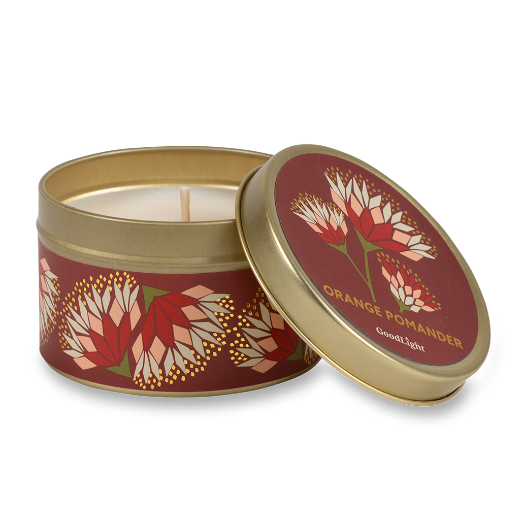 A round metal candle tin with the lid off, showing a white wax candle inside. The tin and lid feature a deep red background with an orange and cream floral pattern and gold lettering that reads "Orange Pomander" with GoodLight branding.