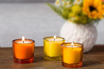 Three votive candles in hues of yellow and orange on a wood table in front of a white vase of flowers.