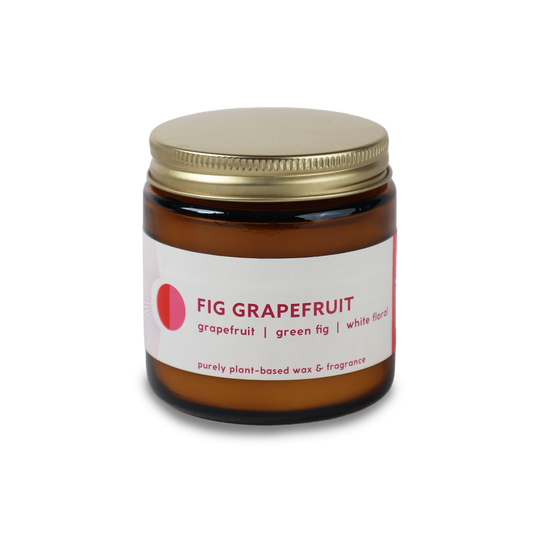 An amber glass jar candle with a gold metal lid and a white label reading 'FIG GRAPEFRUIT' with pink accents, shown on a plain white background.