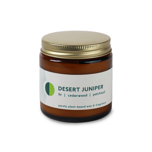 An amber glass jar candle with a gold metal lid and a white label reading 'DESERT JUNIPER' with green and dark green text, photographed on a clean white background.