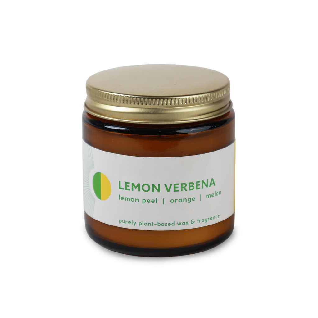 An amber glass jar candle with a gold metal lid and a white label reading 'LEMON VERBENA' with green text and a small yellow-green circular logo, shown on a plain white background.