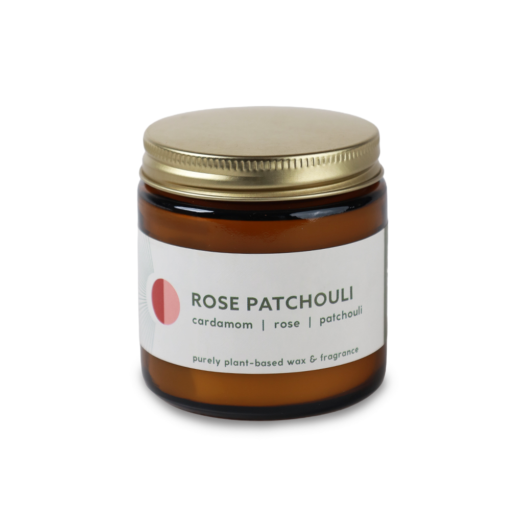 An amber glass jar candle with a gold metal lid and a white label reading 'ROSE PATCHOULI' with 'cardamom | rose | patchouli' underneath, on a plain white background.