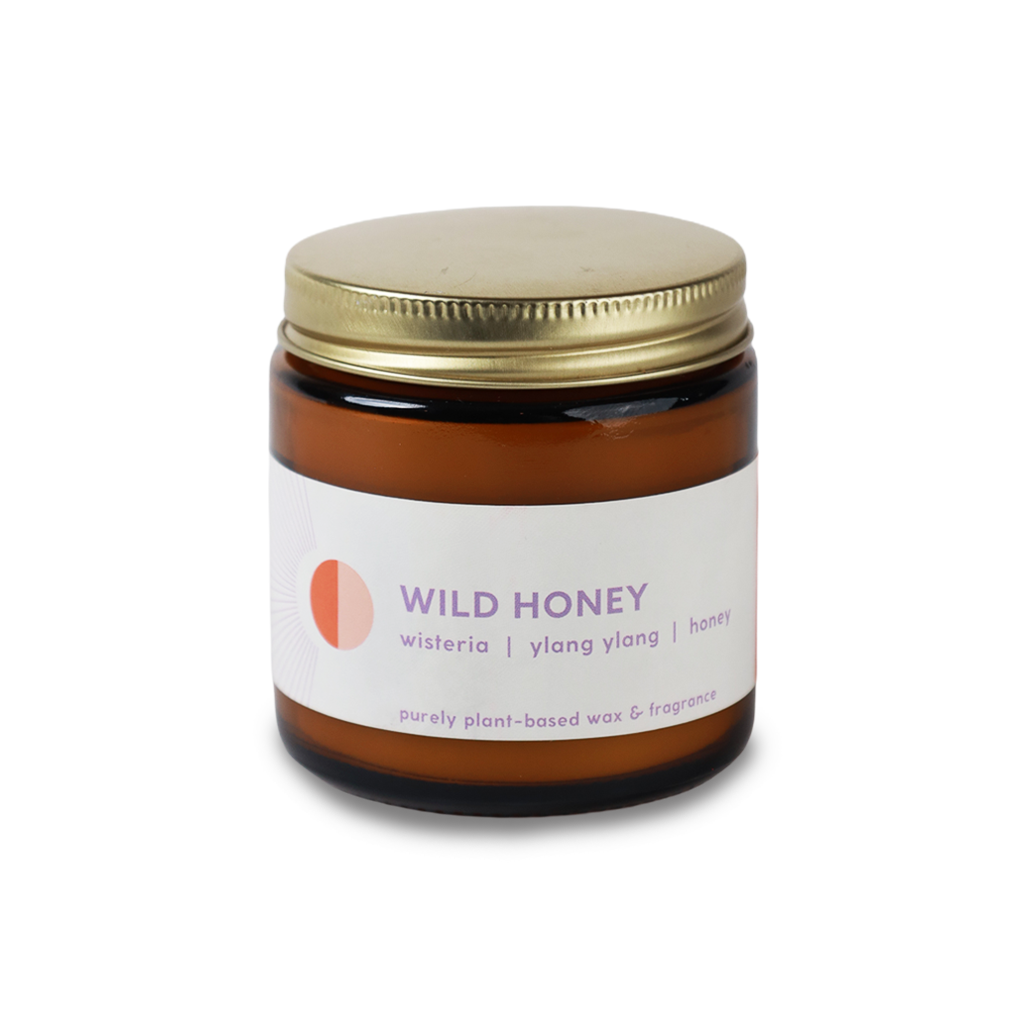 An amber glass jar candle with a gold metal screw-on lid and a white paper label. The label features the text 'WILD HONEY' in purple, along with 'wisteria | ylang ylang | honey' and 'purely plant-based wax & fragrance'.