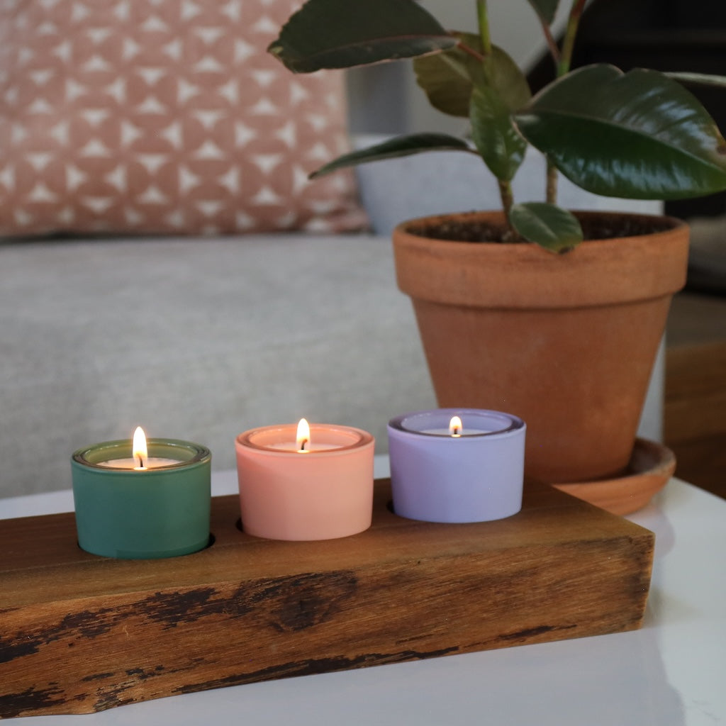 Three colorful votives on a wooden surface with a potted plant in the background.