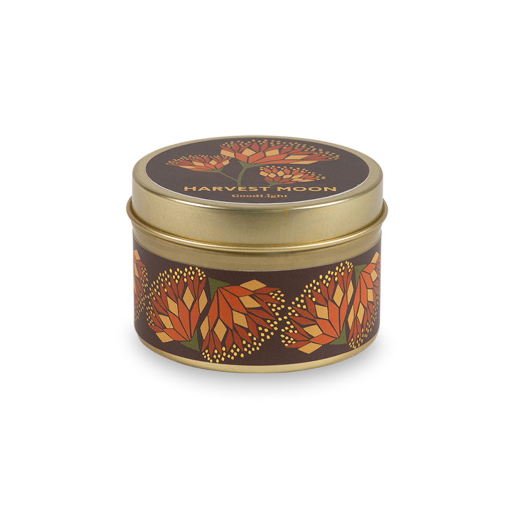 A round metal gold tin candle with a gold lid and base, featuring an autumnal label decorated with orange and pink flowers. The text on the lid reads "Harvest Moon"  with GoodLight branding. 