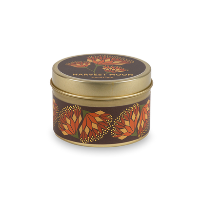 A round metal gold tin candle with a gold lid and base, featuring an autumnal label decorated with orange and pink flowers. The text on the lid reads "Harvest Moon"  with GoodLight branding. 