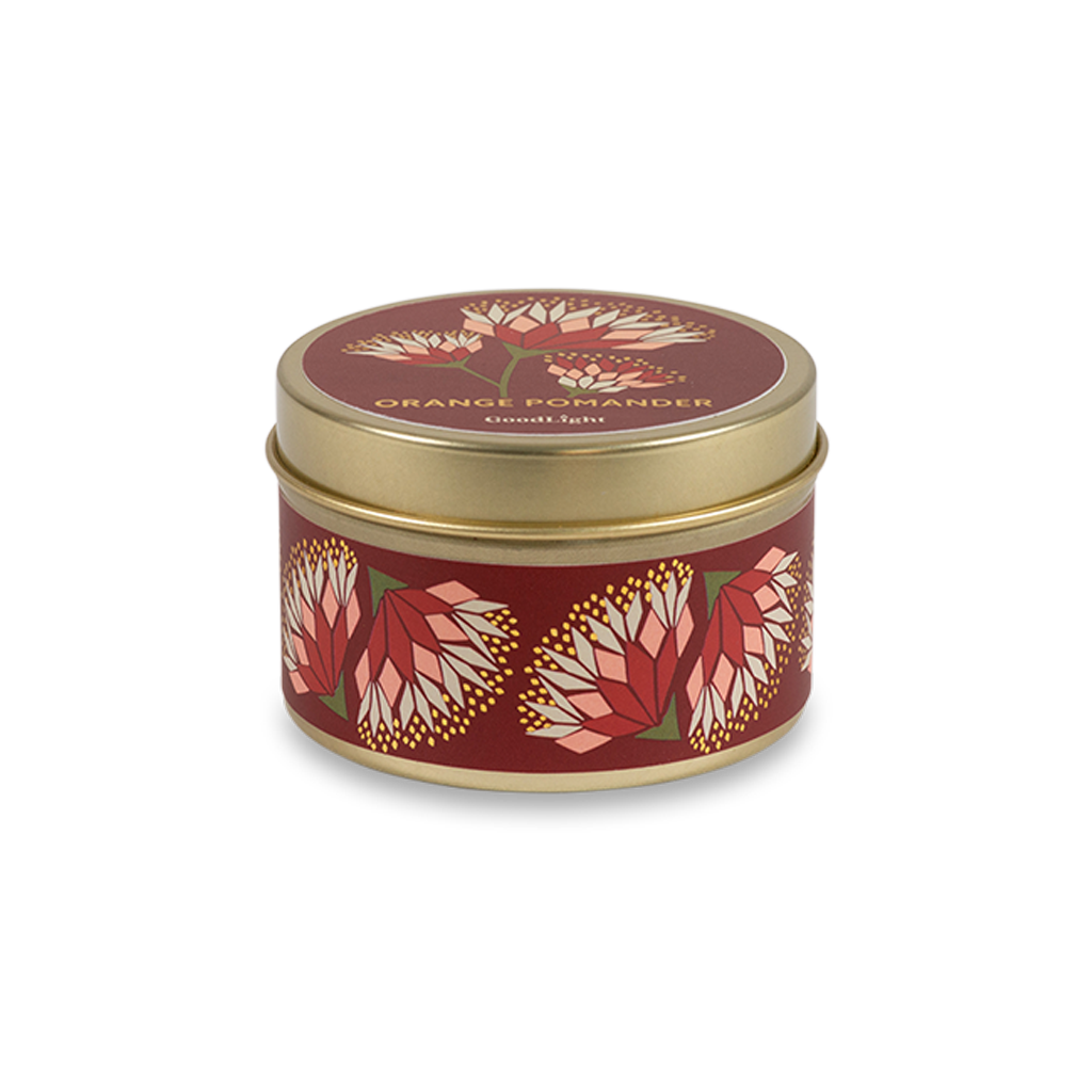 A round metal gold tin candle with a gold lid and base, featuring a deep red label decorated with orange and pink protea flowers. The text on the lid reads "Orange Pomander"  with GoodLight branding. 