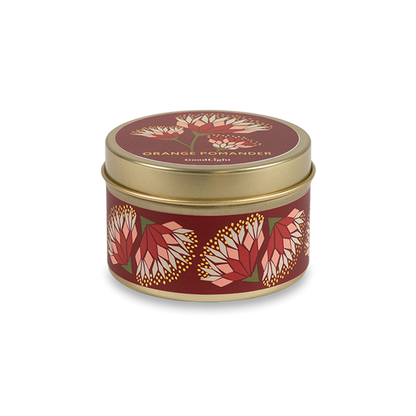 A round metal gold tin candle with a gold lid and base, featuring a deep red label decorated with orange and pink protea flowers. The text on the lid reads "Orange Pomander"  with GoodLight branding. 