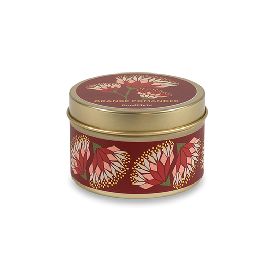 A round metal gold tin candle with a gold lid and base, featuring a deep red label decorated with orange and pink protea flowers. The text on the lid reads "Orange Pomander"  with GoodLight branding. 