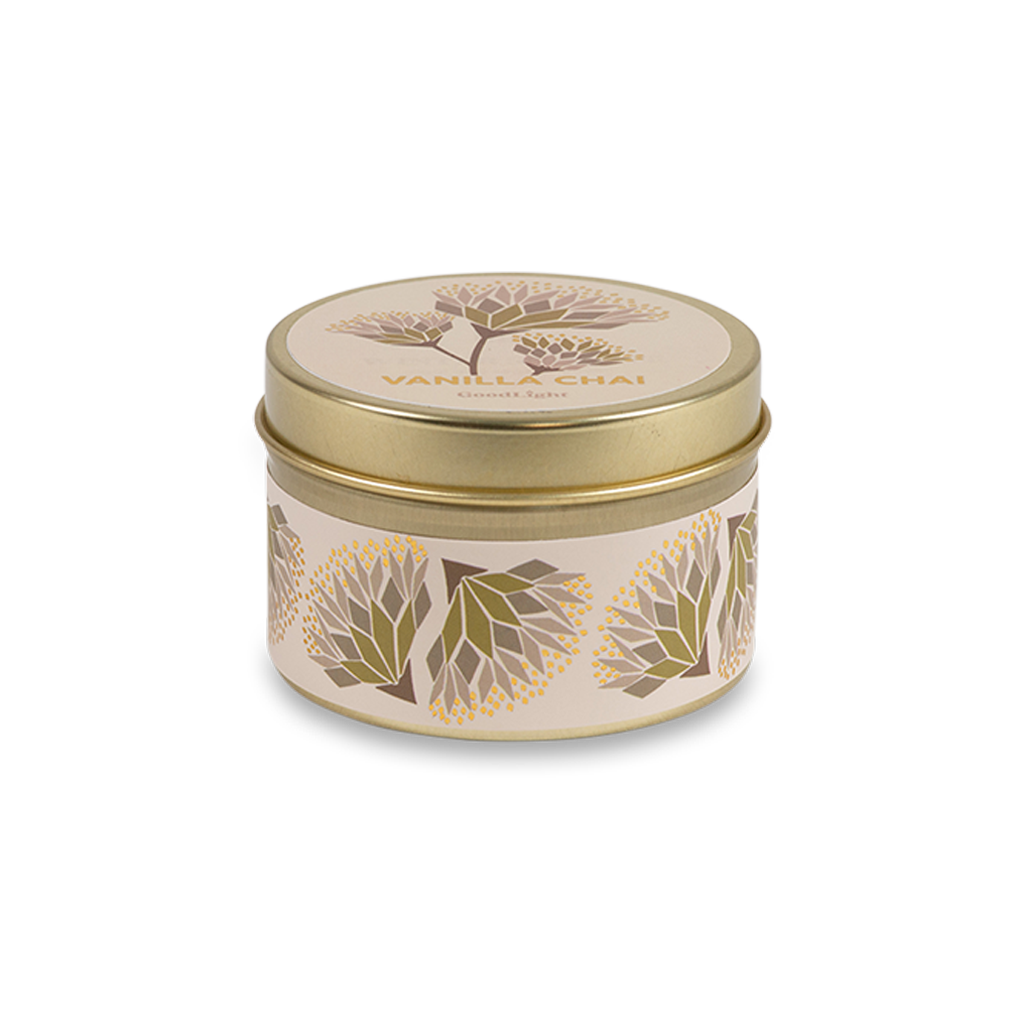 A round metal gold tin candle with a gold lid and base, featuring a cream label decorated with green, purple, and white flowers. The text on the lid reads "Vanilla Chai"  with GoodLight branding. 