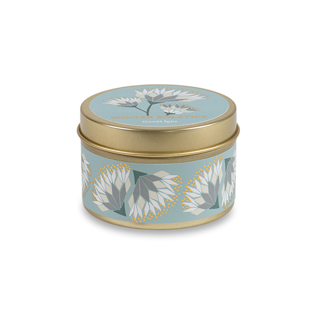 A round metal gold tin candle with a gold lid and base, featuring a light blue label decorated with cream and white flowers. The text on the lid reads "Winter Solstice"  with GoodLight branding. 