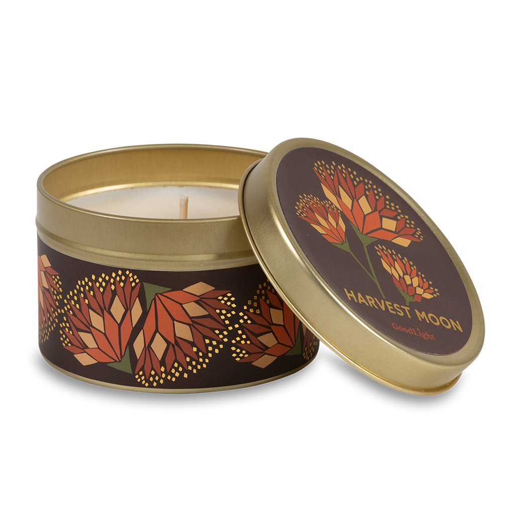 A round metal gold tin candle with a gold lid and base, featuring an autumnal label decorated with orange and pink flowers. The text on the lid reads "Harvest Moon"  with GoodLight branding. 