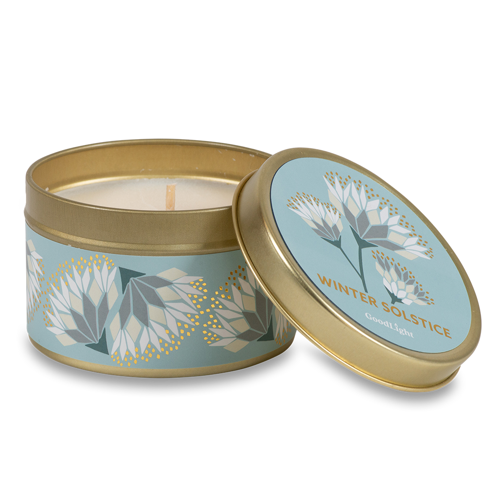 A round metal gold tin candle with a gold lid and base, featuring a light blue label decorated with cream and white flowers. The text on the lid reads "Winter Solstice"  with GoodLight branding. 