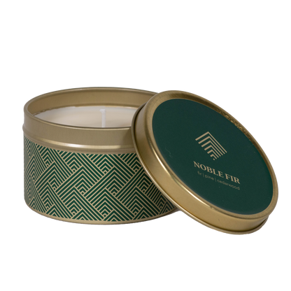 A round metal candle tin with its lid leaning against the base, showing a white candle inside. The tin has a dark green and gold geometric pattern on the body, and the lid features a dark green top with gold text reading "Noble Fire."