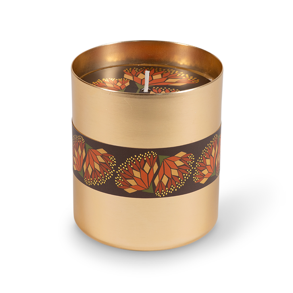 A cylindrical candle in a brushed gold metal container with a dark brown band wrapped around the middle, decorated with a repeating geometric flower pattern in orange, red, and yellow. The candle is shown on a clean white background with a soft shadow.