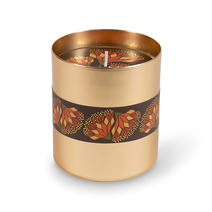 A cylindrical candle in a brushed gold metal container with a dark brown band wrapped around the middle, decorated with a repeating geometric flower pattern in orange, red, and yellow. The candle is shown on a clean white background with a soft shadow.