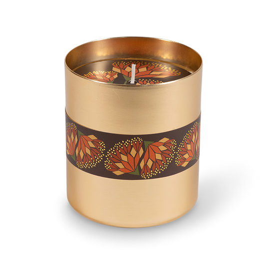 A cylindrical candle in a brushed gold metal container with a dark brown band wrapped around the middle, decorated with a repeating geometric flower pattern in orange, red, and yellow. The candle is shown on a clean white background with a soft shadow.