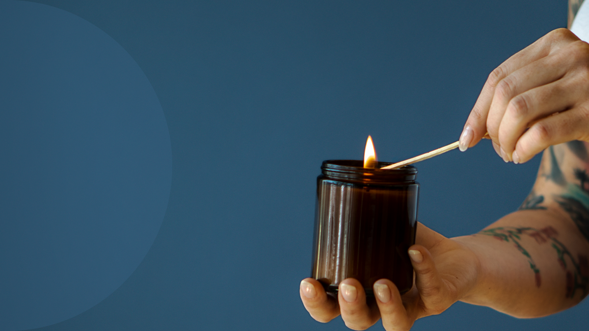 A person's hands holding a dark amber glass jar candle and lighting it with a wooden match against a solid blue background.