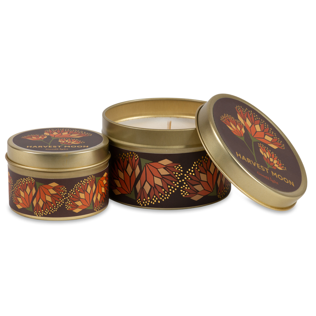Two round metal tins with gold-toned rims and lids, decorated with a dark brown and orange autumn leaf pattern. One tin is open, showing a white wax candle inside, while the other is closed. 