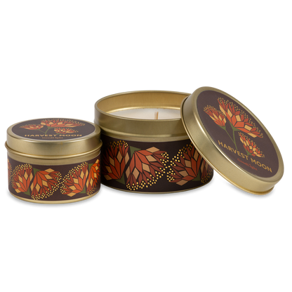 Two round metal tins with gold-toned rims and lids, decorated with a dark brown and orange autumn leaf pattern. One tin is open, showing a white wax candle inside, while the other is closed. 