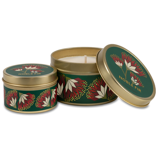 Two round metal tin candles with a festive green, red, and white leaf pattern and gold trim, shown on a clean white background. One tin is open, revealing the white wax inside, while the other one is closed.