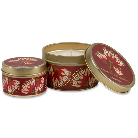 Two round metal candle tins with gold rims and a deep red label featuring orange and cream floral motifs. One tin is open, showing the white wax and wick, while the other is closed. 