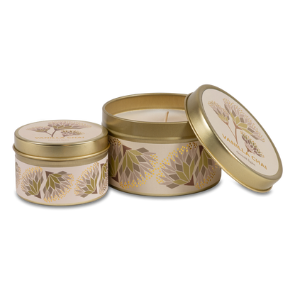 Two round metal candle tins with a gold finish and cream labels featuring botanical illustrations, set against a plain white background.