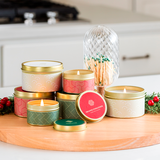Assorted candles in tin containers with Orange Pomander branding on a wooden surface with decorative elements.
